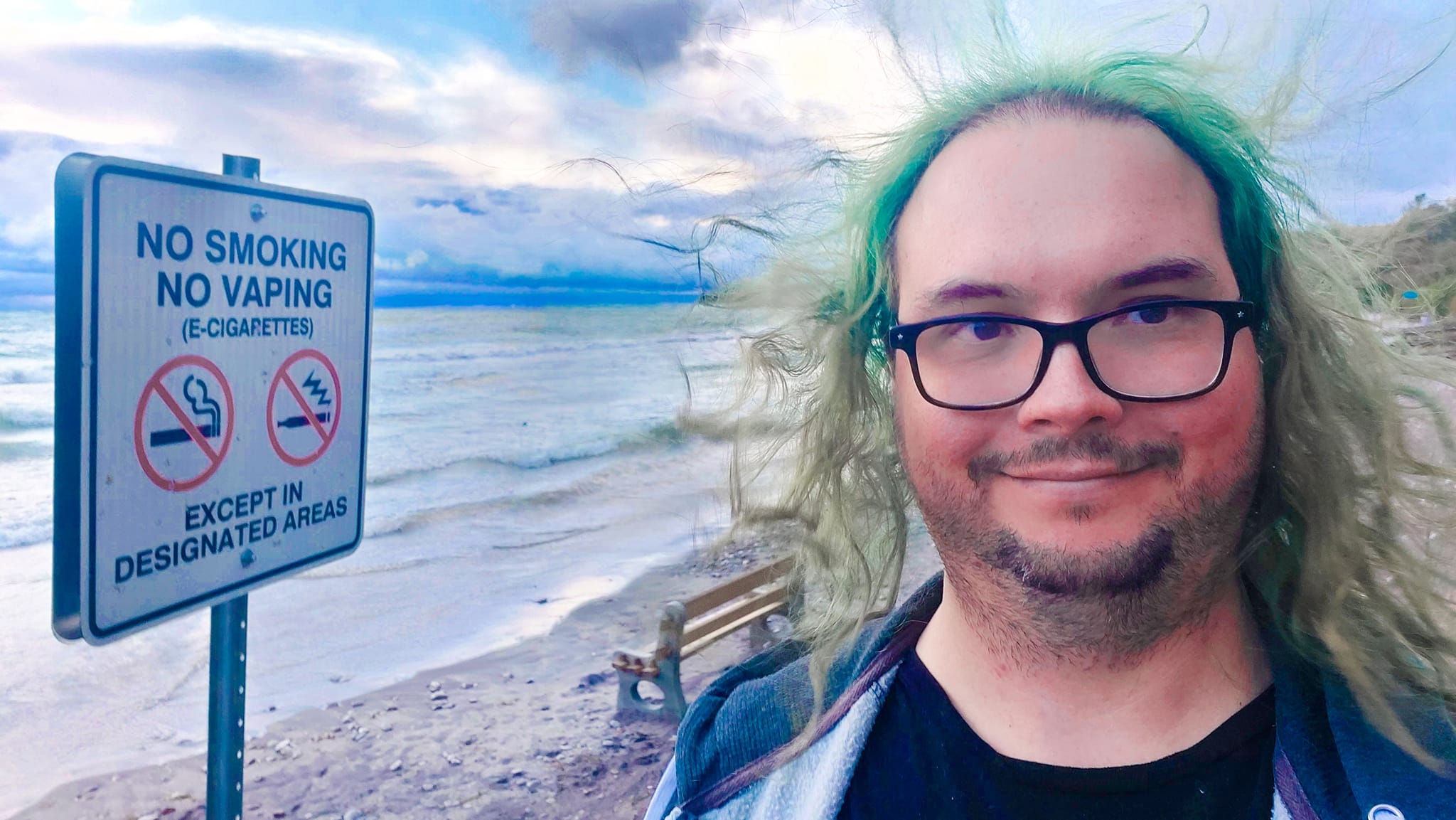 A man with long green hair wearing a hooded sweater vest is on a beach next to a no smoking sign